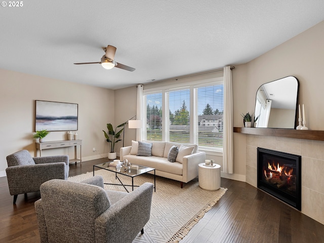 living area featuring baseboards, ceiling fan, dark wood-style flooring, and a tiled fireplace