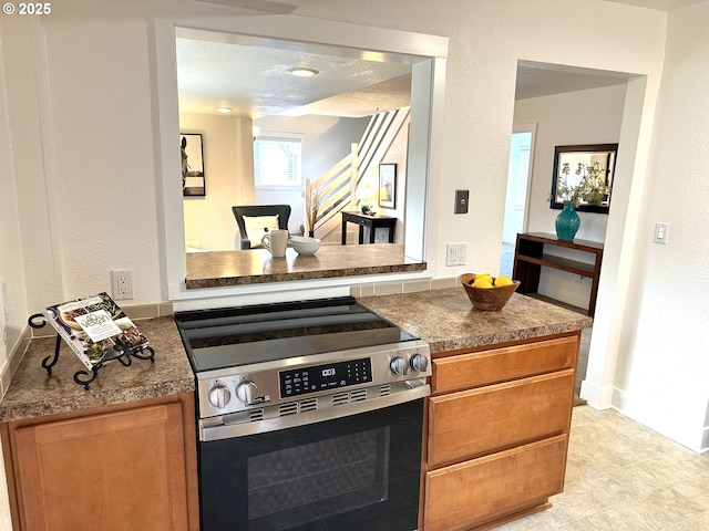 kitchen featuring dark countertops, a textured wall, baseboards, brown cabinetry, and stainless steel range with electric stovetop