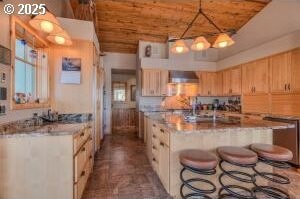 kitchen with wooden ceiling, a kitchen breakfast bar, wall chimney exhaust hood, light brown cabinetry, and a center island with sink