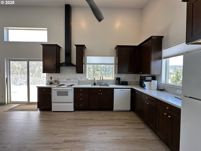 kitchen featuring a towering ceiling, dark brown cabinets, a sink, white appliances, and wall chimney exhaust hood
