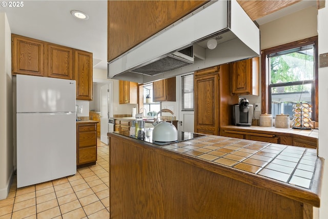 kitchen featuring freestanding refrigerator, tile counters, under cabinet range hood, brown cabinetry, and light tile patterned flooring