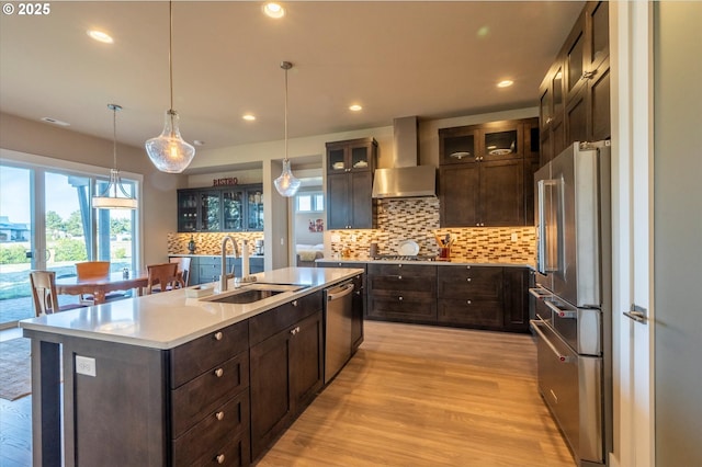 kitchen with appliances with stainless steel finishes, decorative light fixtures, an island with sink, sink, and wall chimney range hood