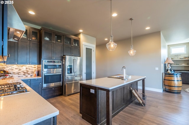 kitchen with appliances with stainless steel finishes, a kitchen island with sink, range hood, dark brown cabinetry, and decorative light fixtures