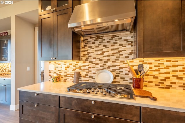 kitchen with dark brown cabinetry, wood-type flooring, stainless steel gas stovetop, decorative backsplash, and exhaust hood