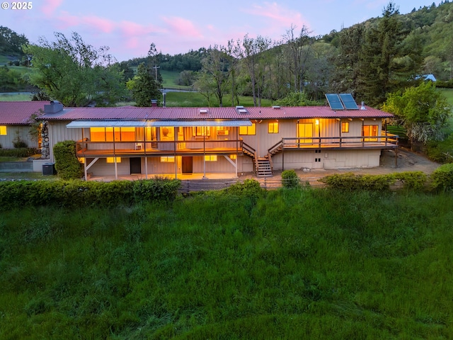 back of property at dusk with board and batten siding, stairway, roof mounted solar panels, a view of trees, and a chimney