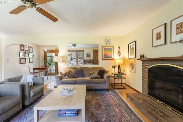 living area featuring a brick fireplace, ceiling fan, baseboards, wood finished floors, and arched walkways