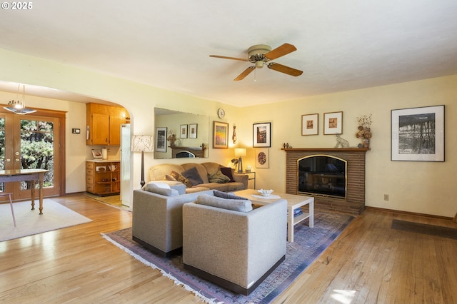 living room featuring light wood-style flooring, a ceiling fan, arched walkways, baseboards, and a brick fireplace