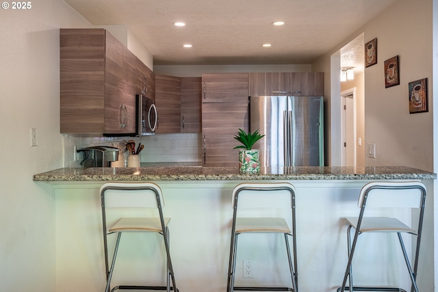 kitchen featuring stone countertops, brown cabinetry, stainless steel appliances, recessed lighting, and decorative backsplash