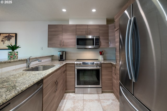 kitchen with appliances with stainless steel finishes, light stone counters, a sink, light tile patterned floors, and backsplash