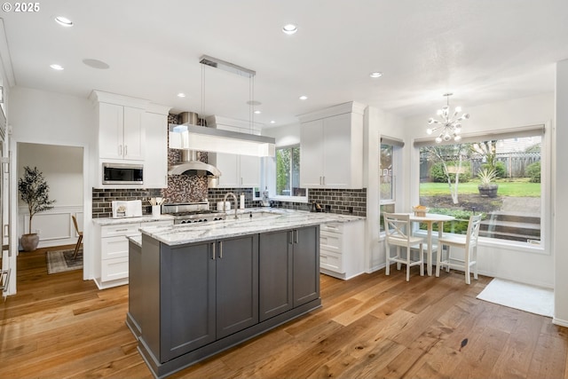 kitchen featuring light wood-type flooring, a kitchen island with sink, stainless steel microwave, tasteful backsplash, and white cabinetry