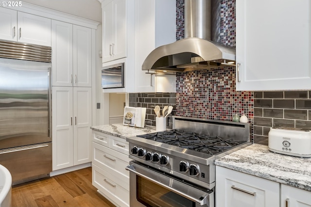 kitchen featuring backsplash, high quality appliances, light wood-style floors, white cabinetry, and wall chimney exhaust hood