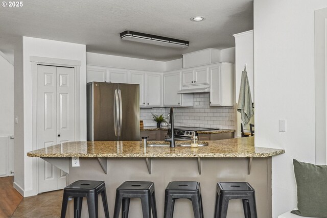 kitchen featuring freestanding refrigerator, under cabinet range hood, a peninsula, light stone countertops, and recessed lighting