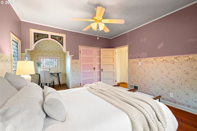 bedroom with a wainscoted wall, wood finished floors, visible vents, ornamental molding, and wallpapered walls