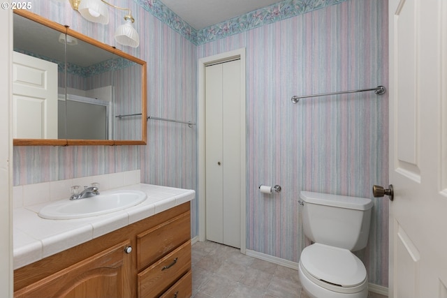 bathroom with a shower with shower door, toilet, vanity, baseboards, and wallpapered walls