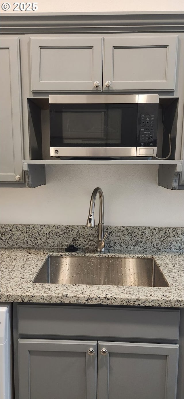 kitchen view of a sink, gray cabinets, stainless steel microwave, light stone counters, and open shelves