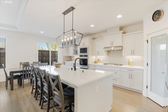 kitchen featuring decorative backsplash, stainless steel appliances, under cabinet range hood, light countertops, and a center island with sink
