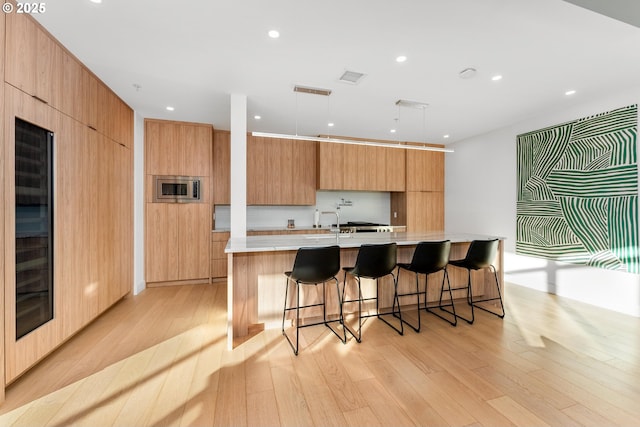 kitchen with a spacious island, hanging light fixtures, stainless steel microwave, a kitchen breakfast bar, and light hardwood / wood-style floors