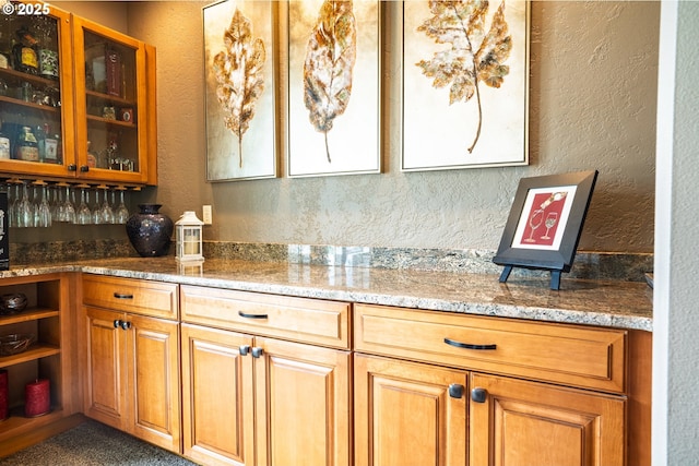 kitchen featuring a textured wall, light stone countertops, glass insert cabinets, brown cabinetry, and open shelves
