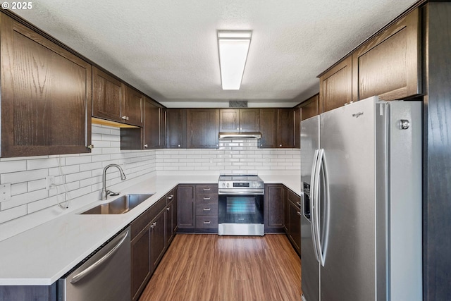 kitchen featuring wood finished floors, dark brown cabinetry, under cabinet range hood, a sink, and appliances with stainless steel finishes