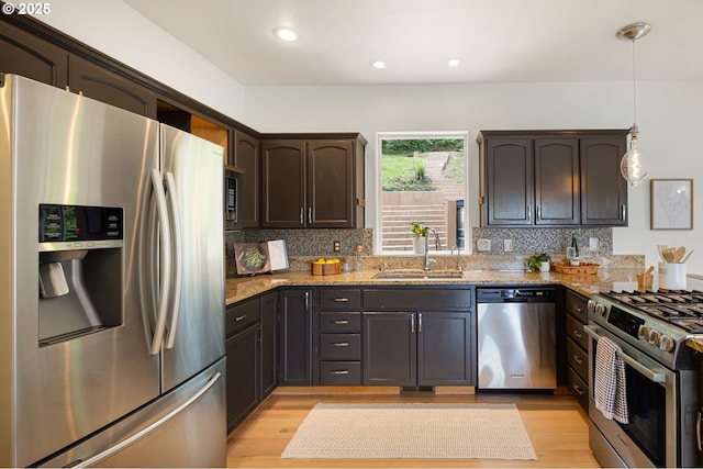 kitchen with appliances with stainless steel finishes, a sink, dark brown cabinetry, light stone countertops, and light wood-style flooring