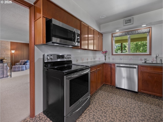 kitchen featuring dark colored carpet, wooden walls, brown cabinets, a sink, and appliances with stainless steel finishes