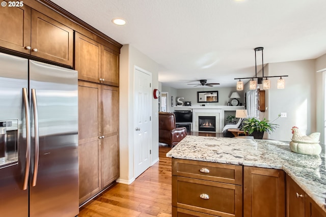 kitchen with stainless steel fridge with ice dispenser, a glass covered fireplace, light stone counters, light wood-style flooring, and ceiling fan