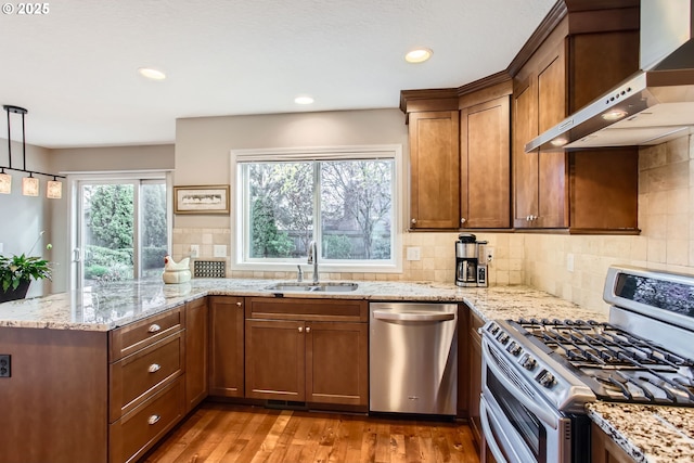 kitchen featuring wall chimney exhaust hood, a sink, appliances with stainless steel finishes, brown cabinetry, and light wood finished floors