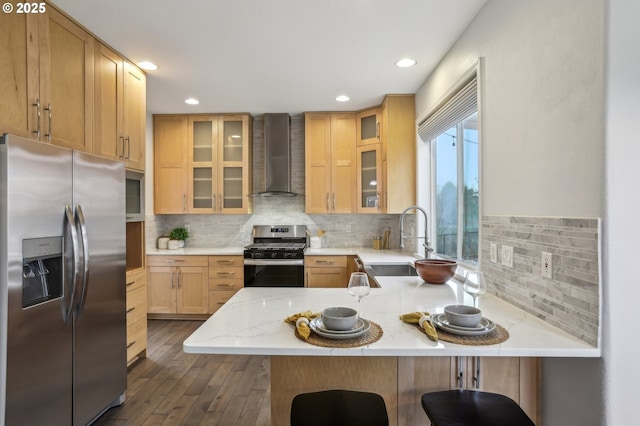 kitchen with a breakfast bar, kitchen peninsula, appliances with stainless steel finishes, light brown cabinetry, and wall chimney exhaust hood