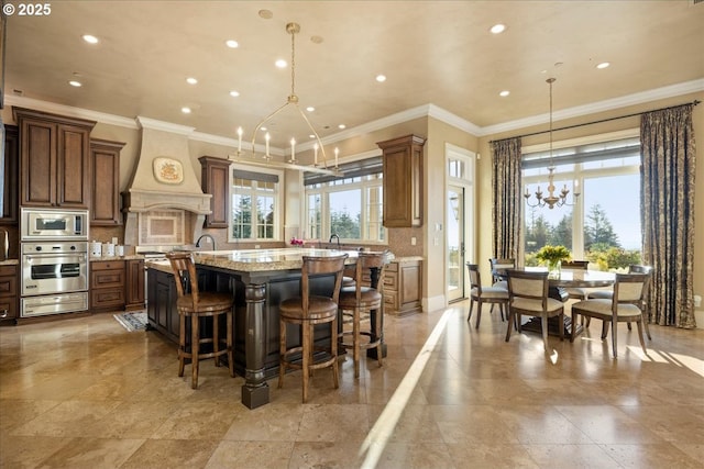 kitchen with premium range hood, a chandelier, a large island with sink, appliances with stainless steel finishes, and light stone countertops