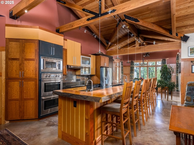 kitchen with concrete flooring, stainless steel appliances, wooden ceiling, vaulted ceiling with beams, and a center island with sink