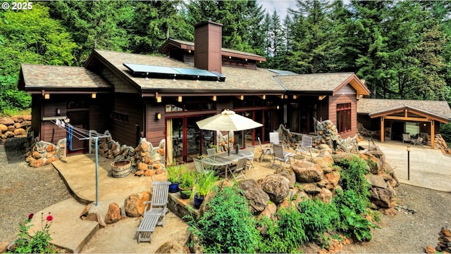 rear view of property featuring outdoor dining area, roof with shingles, a chimney, a patio area, and solar panels