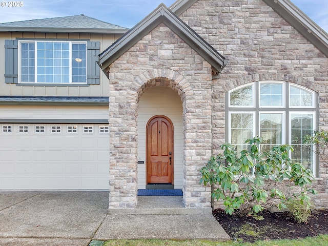 view of exterior entry featuring a garage, a shingled roof, driveway, stone siding, and board and batten siding