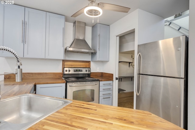 kitchen with a sink, butcher block countertops, wall chimney exhaust hood, a ceiling fan, and appliances with stainless steel finishes