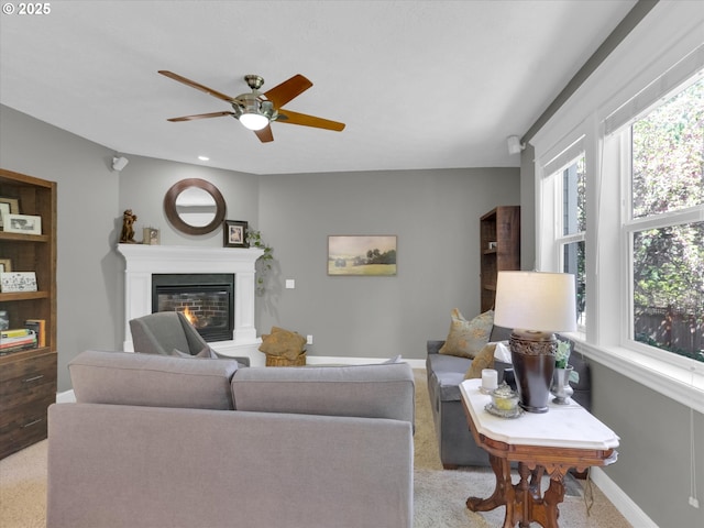living area featuring a ceiling fan, baseboards, a glass covered fireplace, and light carpet