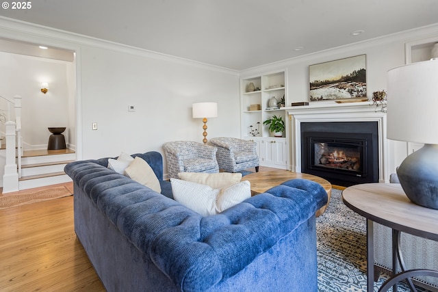 living room featuring light wood finished floors, ornamental molding, a glass covered fireplace, built in features, and stairway