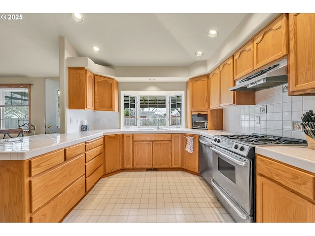 kitchen with a peninsula, tile counters, under cabinet range hood, appliances with stainless steel finishes, and backsplash
