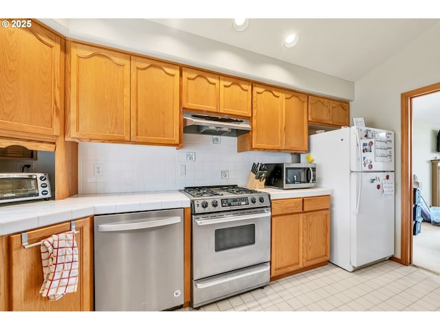 kitchen featuring a toaster, tile countertops, under cabinet range hood, tasteful backsplash, and appliances with stainless steel finishes