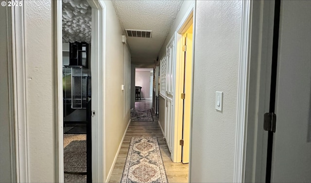 corridor featuring light wood-type flooring, visible vents, a textured ceiling, and a textured wall