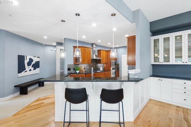3646 SW Baird St Unit 3, Portland, OR 97219 - kitchen with stainless steel appliances, wall chimney range hood, brown cabinetry, decorative backsplash, and light wood-style flooring
