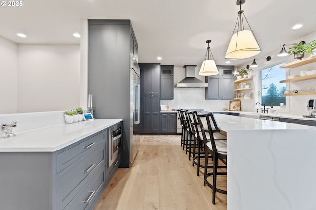 kitchen with a breakfast bar, gray cabinets, wall chimney range hood, open shelves, and light wood finished floors