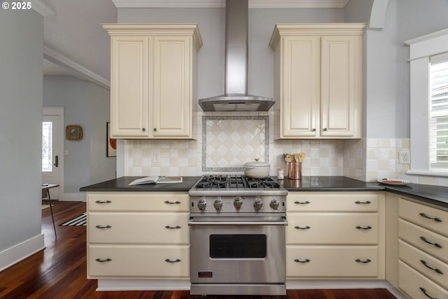 kitchen featuring wall chimney exhaust hood, cream cabinetry, dark countertops, and designer stove