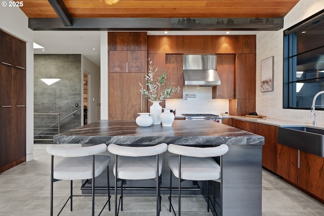 kitchen featuring wall chimney range hood, brown cabinets, beam ceiling, a sink, and modern cabinets