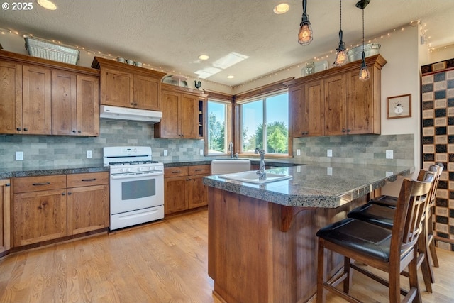 kitchen featuring white gas range, under cabinet range hood, brown cabinets, a kitchen bar, and a peninsula