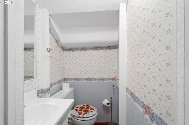 half bathroom featuring vanity, a textured ceiling, toilet, and wallpapered walls