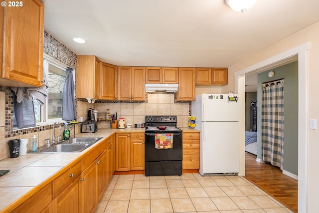 kitchen featuring tile countertops, under cabinet range hood, black range with electric stovetop, a sink, and freestanding refrigerator