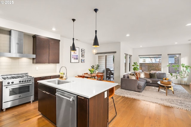 kitchen with a center island with sink, decorative light fixtures, sink, appliances with stainless steel finishes, and wall chimney exhaust hood