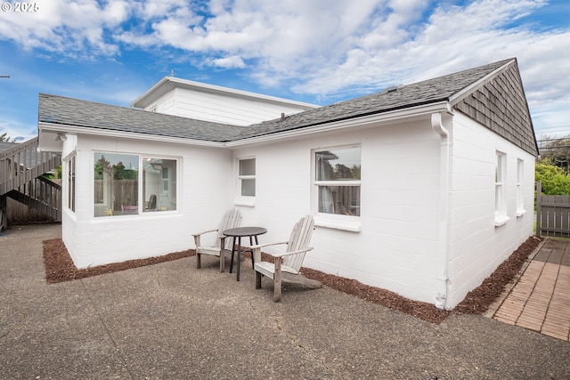 rear view of house with fence, a patio, roof with shingles, and concrete block siding