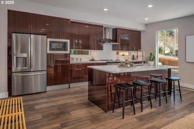 kitchen featuring a kitchen bar, dark hardwood / wood-style floors, stainless steel appliances, a kitchen island with sink, and wall chimney range hood