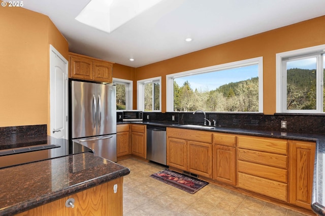 kitchen featuring dark stone counters, a skylight, brown cabinetry, stainless steel appliances, and a sink