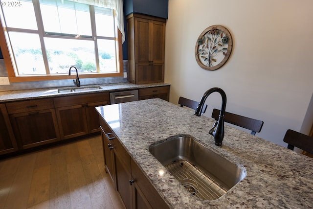 kitchen with stainless steel dishwasher, wood-type flooring, light stone counters, and a sink